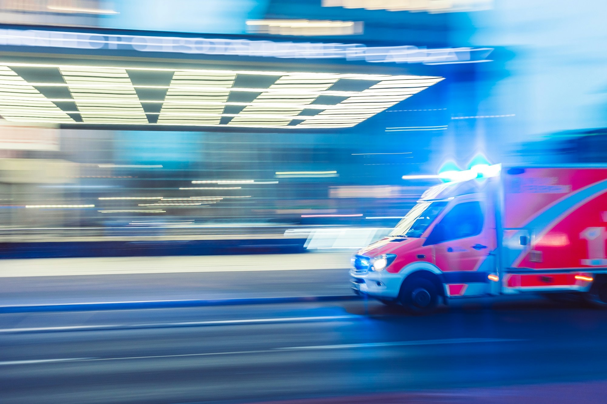An ambulance moving through a blue-lit city at night, its lights streaked with motion